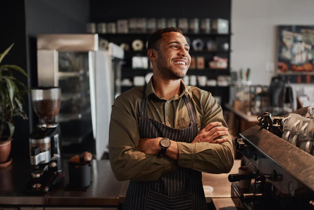 Young afro-american small coffee shop owner standing behind counter wearing apron with crossed arms looking away