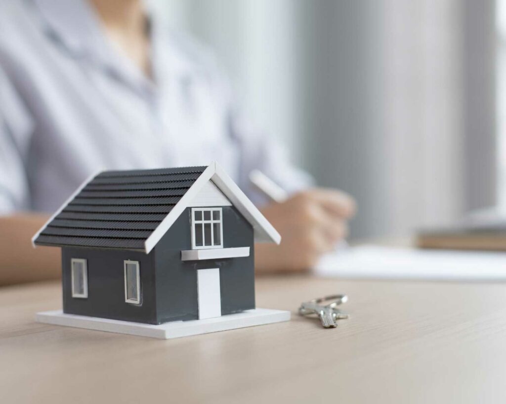 small, fake house in the foreground on a desk with keys, someone writing in the background